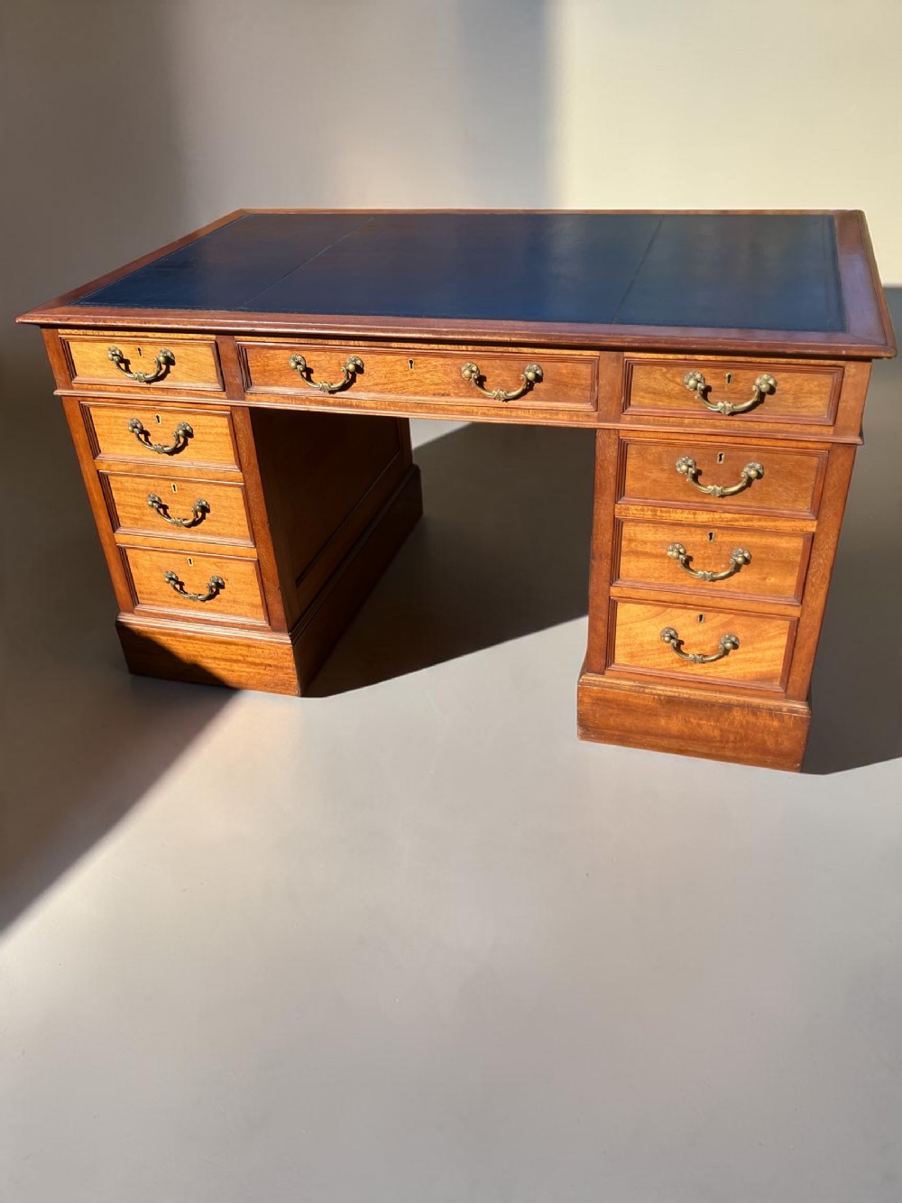A mahogany pedestal desk featuring a dark blue leather writing top, brass swan-neck handles, and nine drawers arranged across two pedestals.