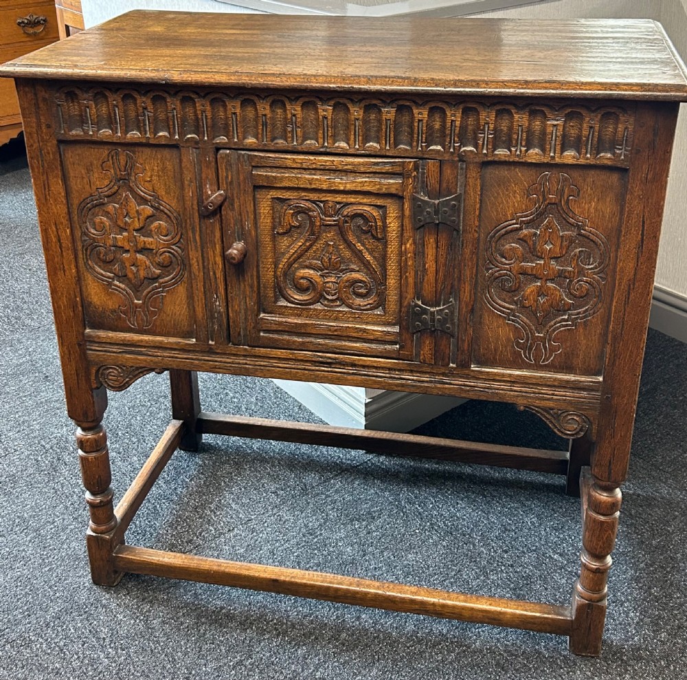 A dark carved oak side cabinet featuring scrollwork panels, an arcaded top frieze, iron strap hinges, turned legs, and a lower stretcher base.