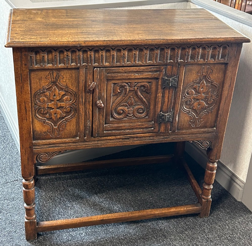 A dark carved oak side cabinet featuring scrollwork panels, an arcaded top frieze, iron strap hinges, turned legs, and a lower stretcher base. View II