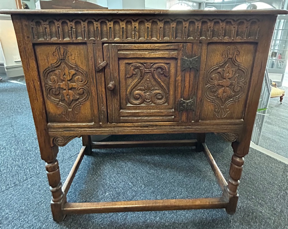 A dark carved oak side cabinet featuring scrollwork panels, an arcaded top frieze, iron strap hinges, turned legs, and a lower stretcher base. View III