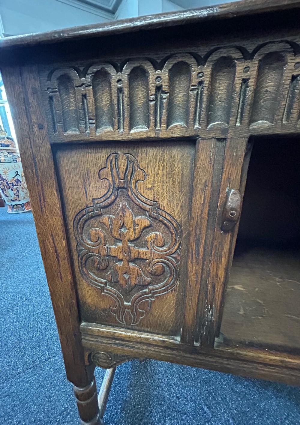 A dark carved oak side cabinet featuring scrollwork panels, an arcaded top frieze, iron strap hinges, turned legs, and a lower stretcher base. View IV