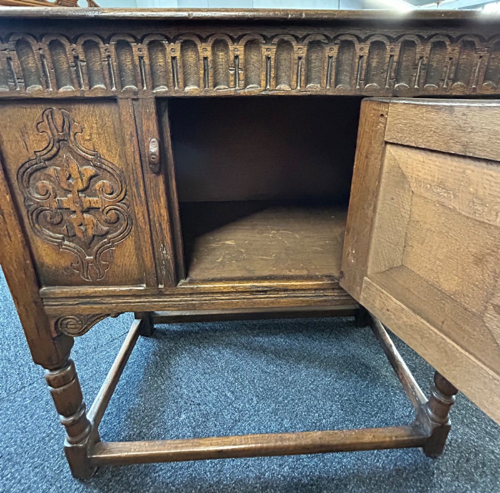 A dark carved oak side cabinet featuring scrollwork panels, an arcaded top frieze, iron strap hinges, turned legs, and a lower stretcher base. View V