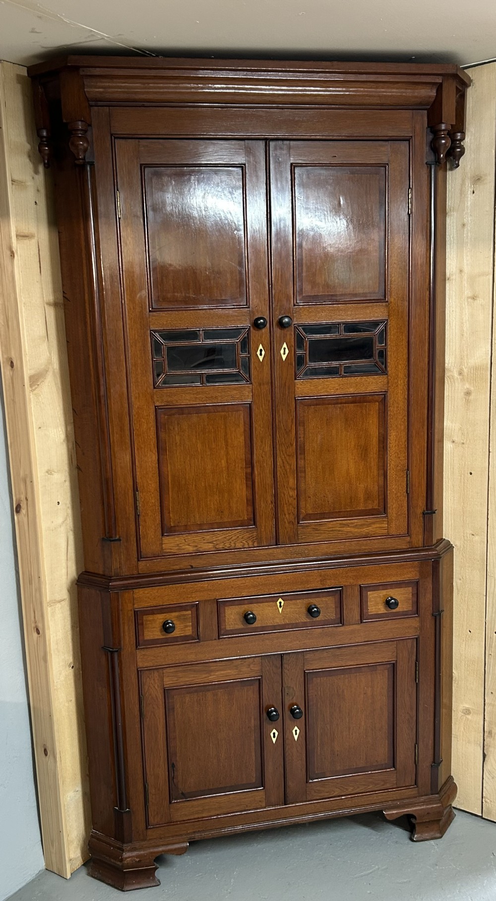 A tall freestanding antique Welsh oak corner cabinet with upper leaded glass doors, turned pendant drop mouldings, middle drawers, and lower panelled doors.