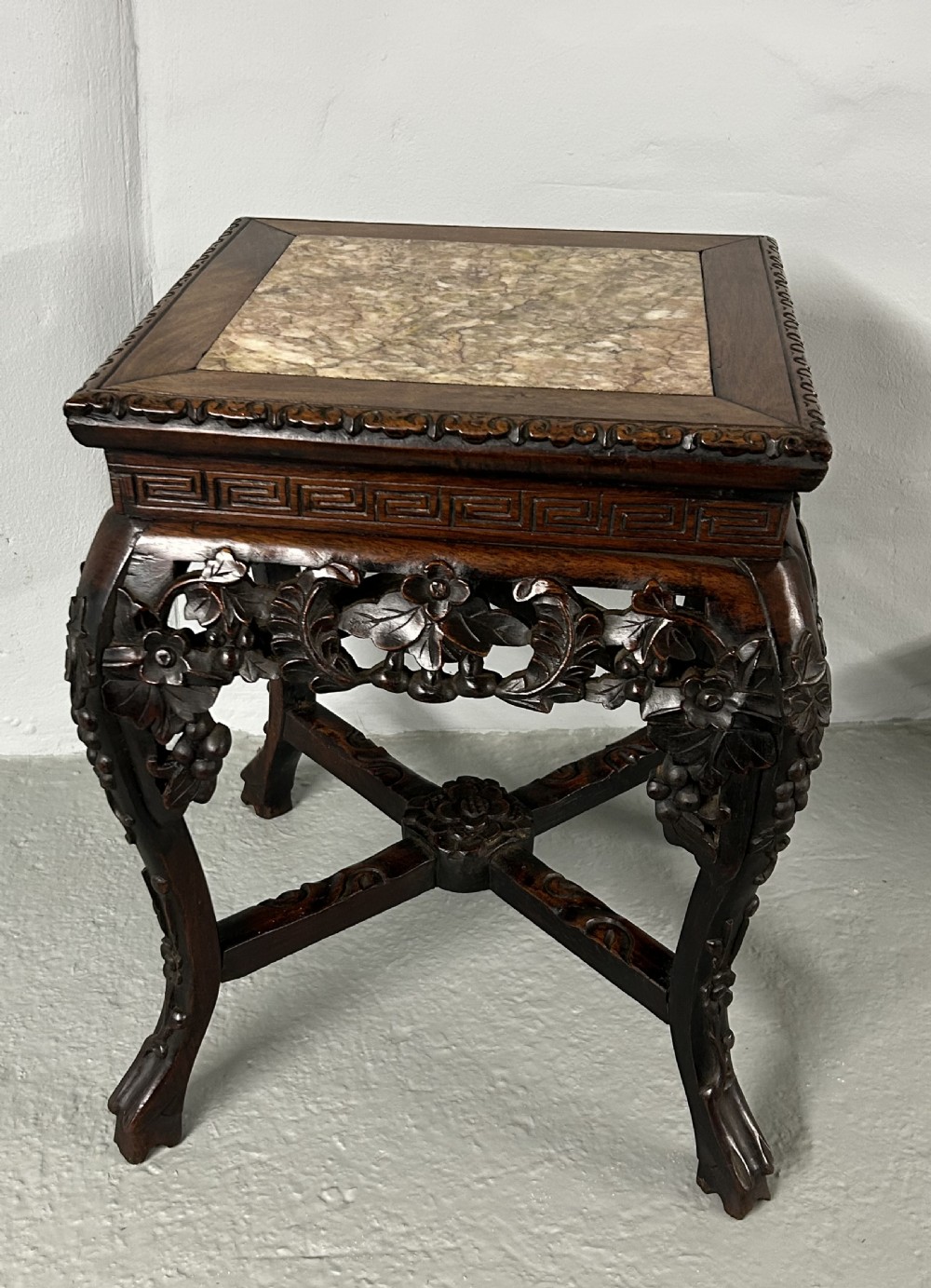 A square carved dark hardwood side table featuring an inset variegated marble top, pierced floral apron, and curved legs with an X-stretcher.