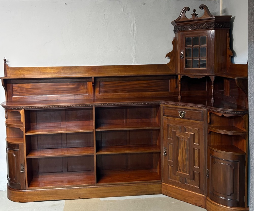 A large Victorian mahogany corner bookcase featuring open shelves along one section, a central panelled door cupboard, and an elevated glazed corner cabinet topped with a swan-neck pediment. View II