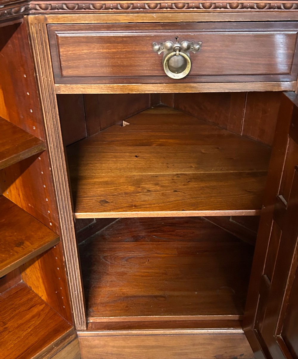 A large Victorian mahogany corner bookcase featuring open shelves along one section, a central panelled door cupboard, and an elevated glazed corner cabinet topped with a swan-neck pediment. View V