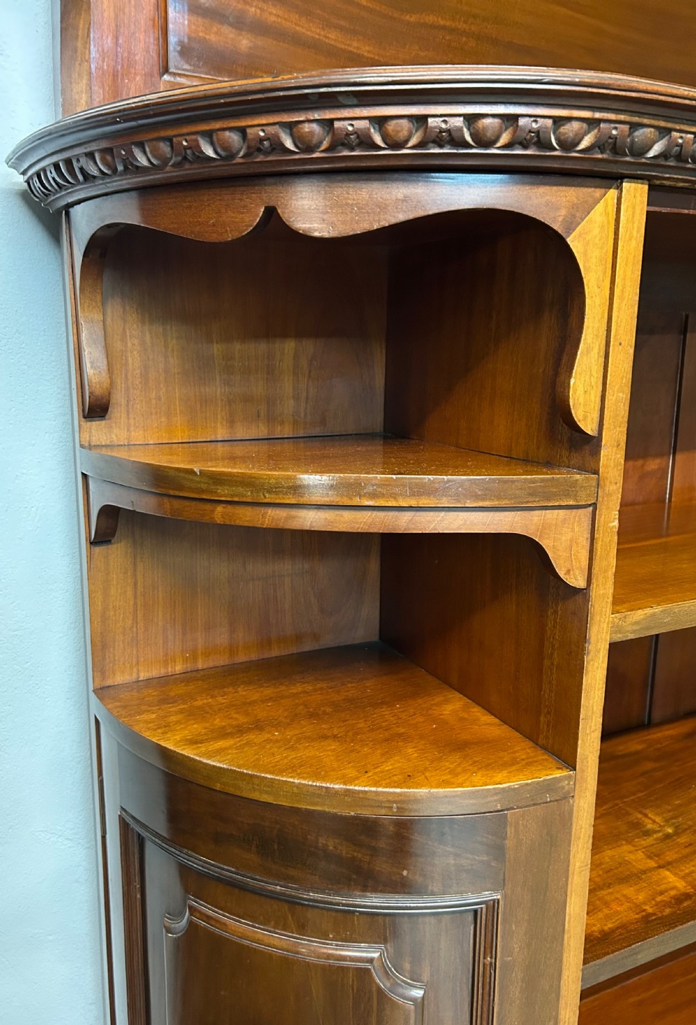 A large Victorian mahogany corner bookcase featuring open shelves along one section, a central panelled door cupboard, and an elevated glazed corner cabinet topped with a swan-neck pediment. View X