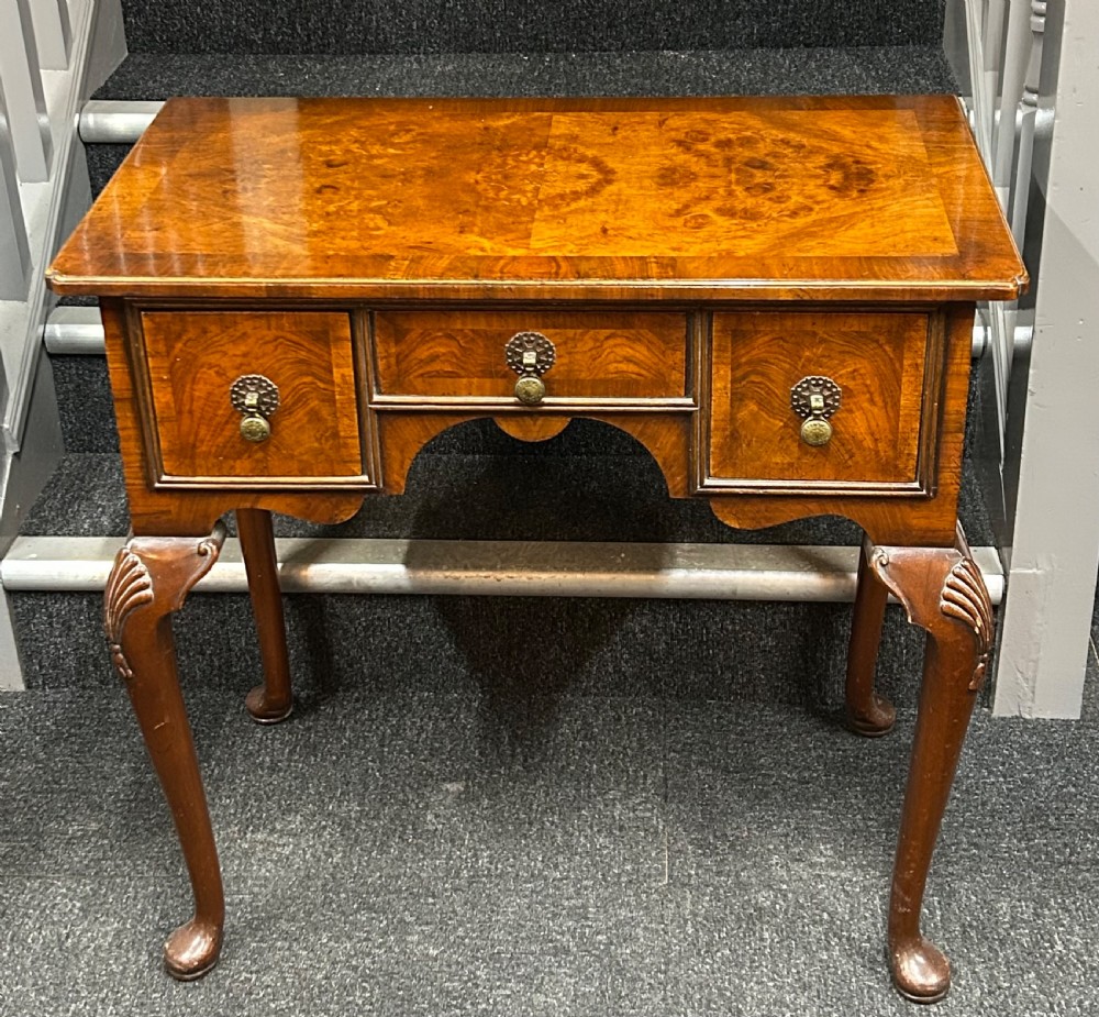 A 1920s Queen Anne style burr walnut lowboy featuring three drawers with brass handles, carved cabriole legs, and pad feet.
