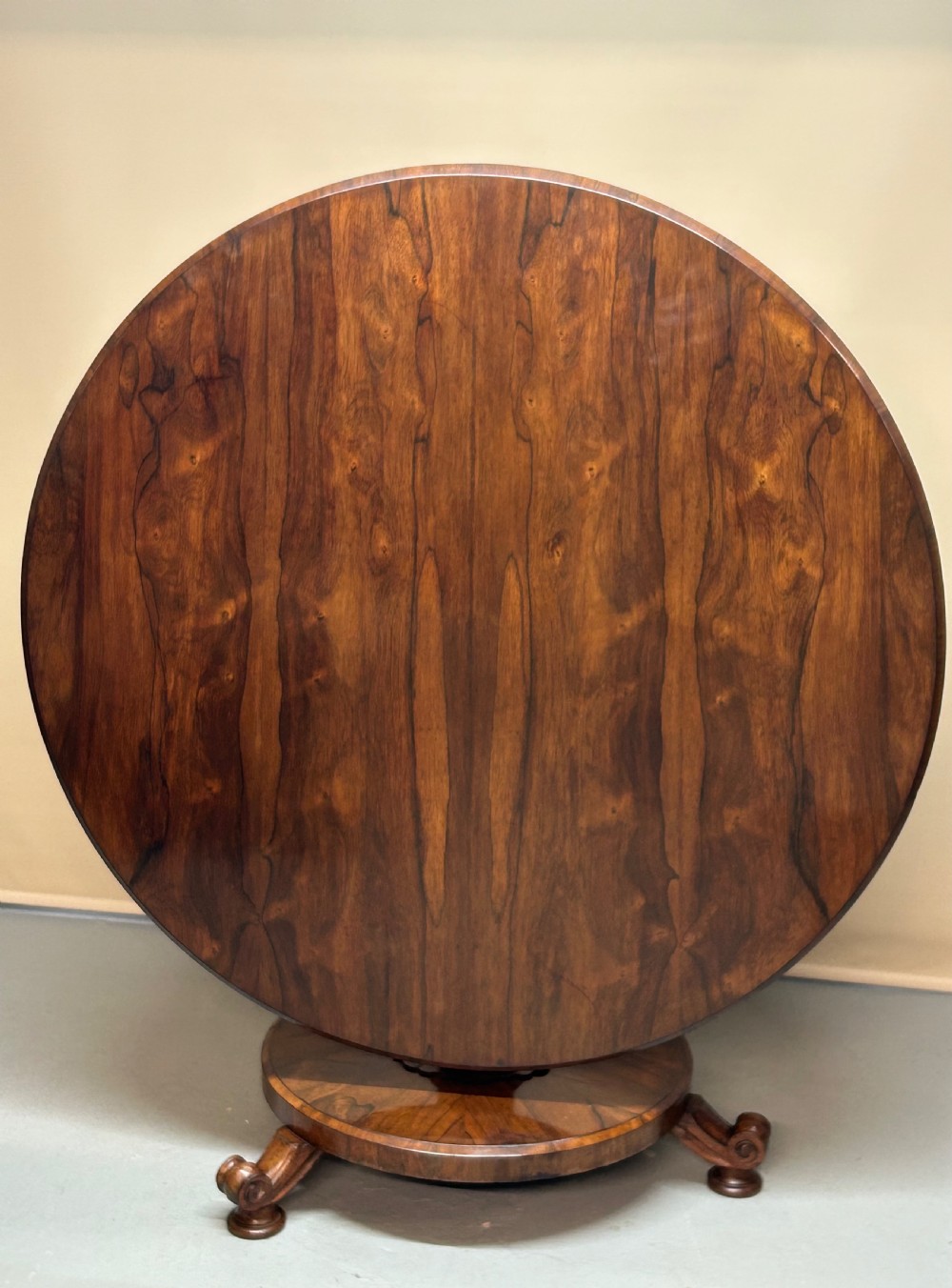 A circular William IV tilt-top rosewood breakfast table featuring dark figured grain, shown upright on a circular plinth base with carved scroll feet.