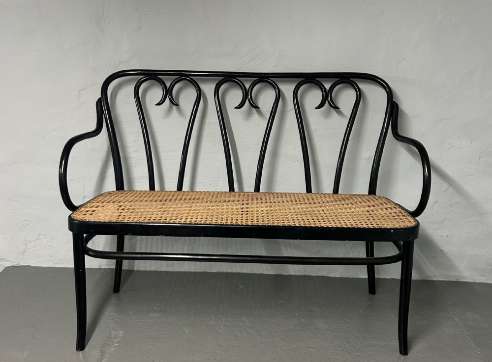 A black painted bentwood two-seater bench featuring three heart-shaped backrest scrolls and a woven natural rattan cane seat.