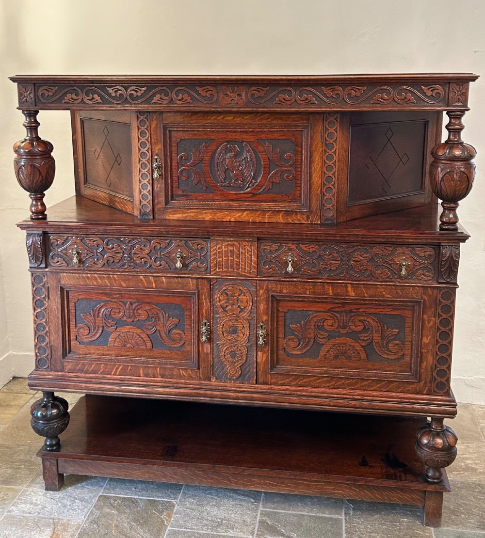 A dark carved oak court cupboard featuring bulbous turned supports, relief scrollwork carvings, a central panel with an eagle motif, and a lower open shelf.