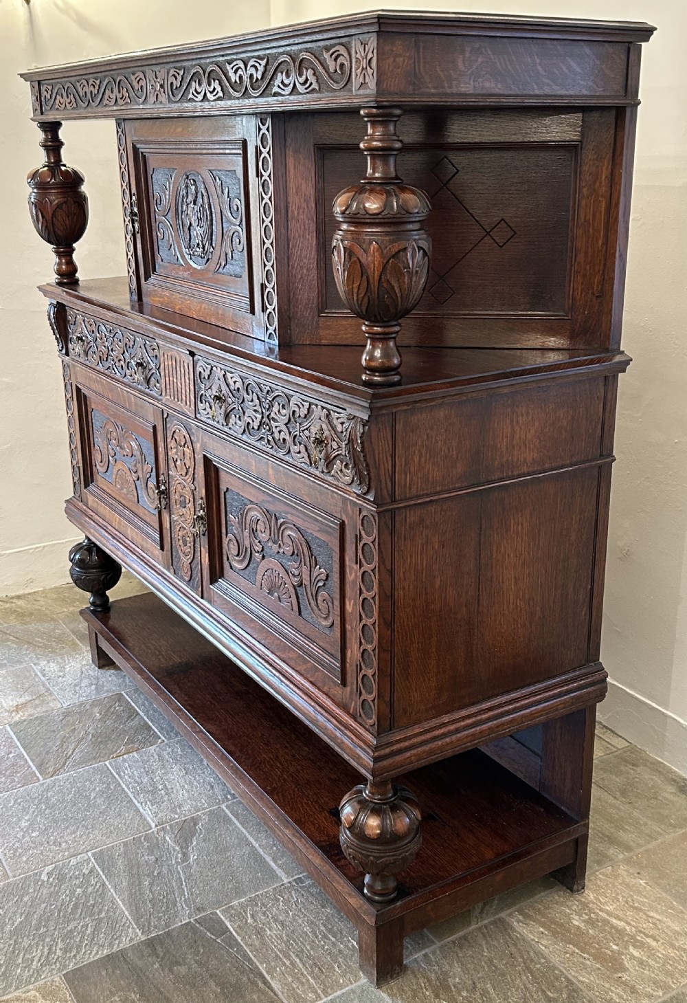 A dark carved oak court cupboard featuring bulbous turned supports, relief scrollwork carvings, a central panel with an eagle motif, and a lower open shelf. View II