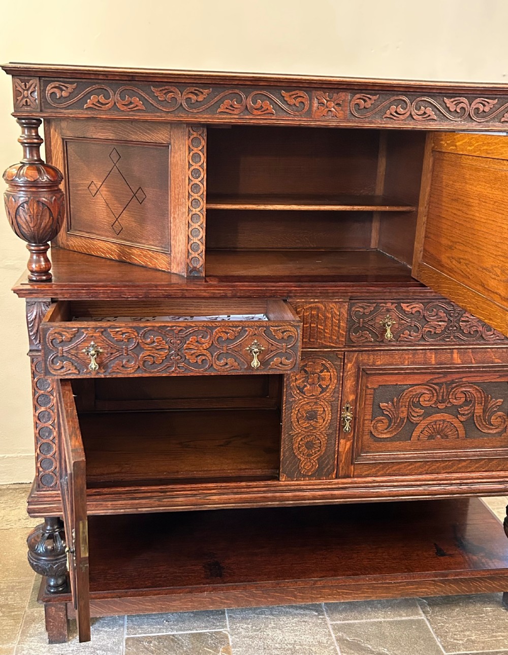 A dark carved oak court cupboard featuring bulbous turned supports, relief scrollwork carvings, a central panel with an eagle motif, and a lower open shelf. View IV
