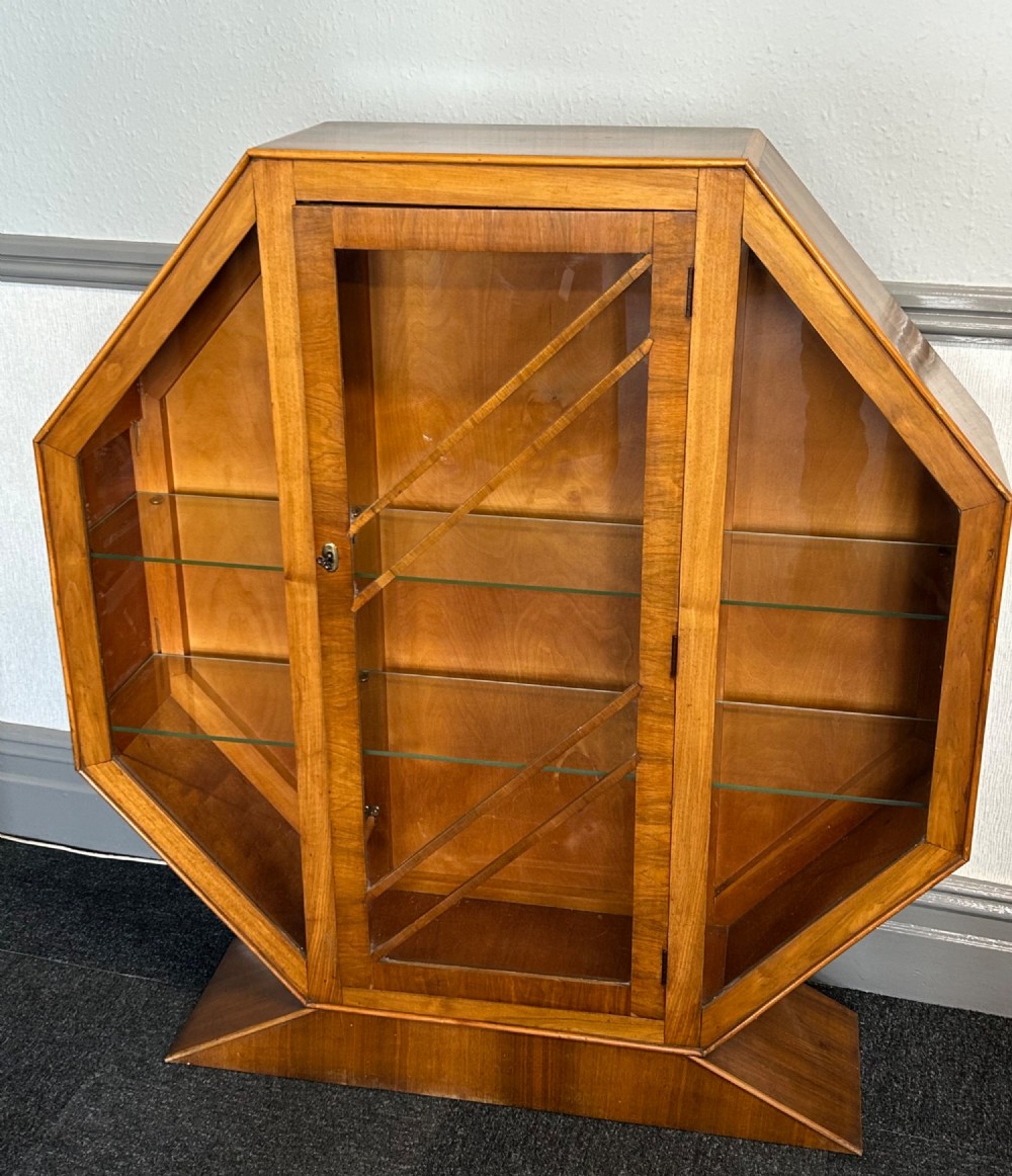 An octagonal Art Deco walnut display cabinet featuring a central glazed door with diagonal glazing bars and glass interior shelves on a flared plinth base.