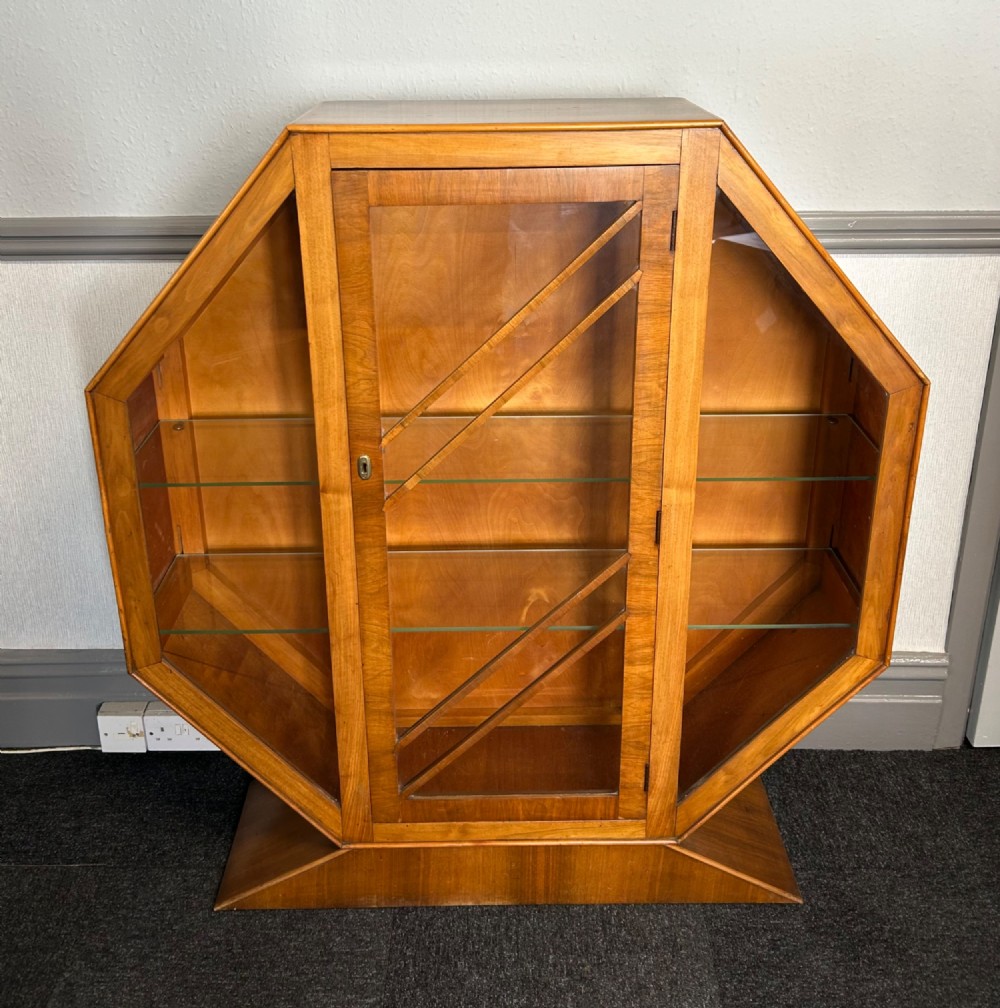 An octagonal Art Deco walnut display cabinet featuring a central glazed door with diagonal glazing bars and glass interior shelves on a flared plinth base. View II
