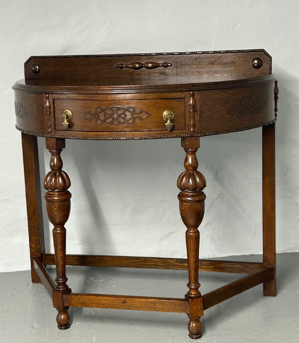 A 1920s dark oak demi-lune console table featuring a carved strapwork drawer, turned front legs, and a low gallery back.