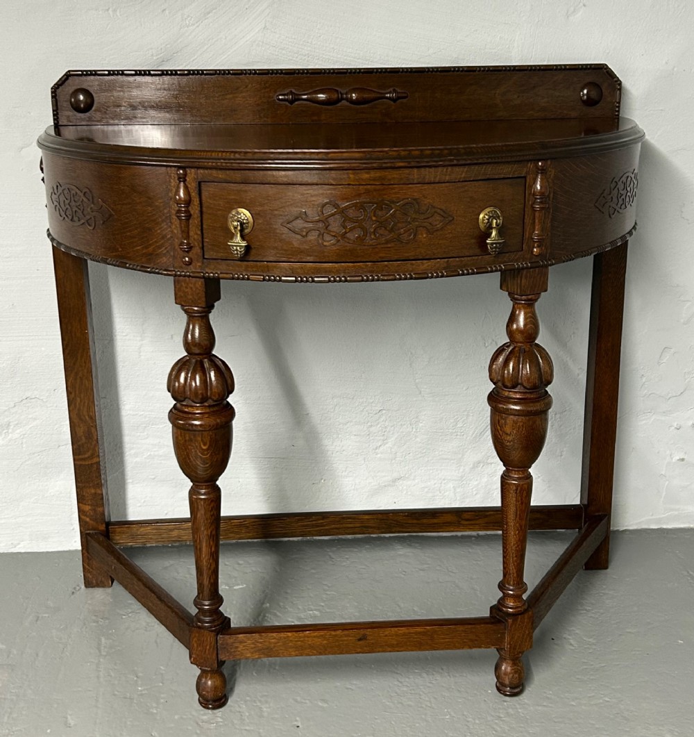 A 1920s dark oak demi-lune console table featuring a carved strapwork drawer, turned front legs, and a low gallery back. View II