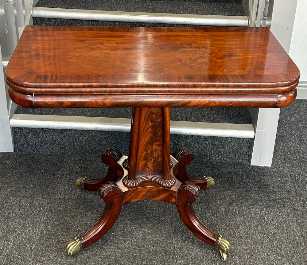 A William IV mahogany fold-over tea table with a flame mahogany top, central carved pedestal, and four splayed legs with brass paw castors.