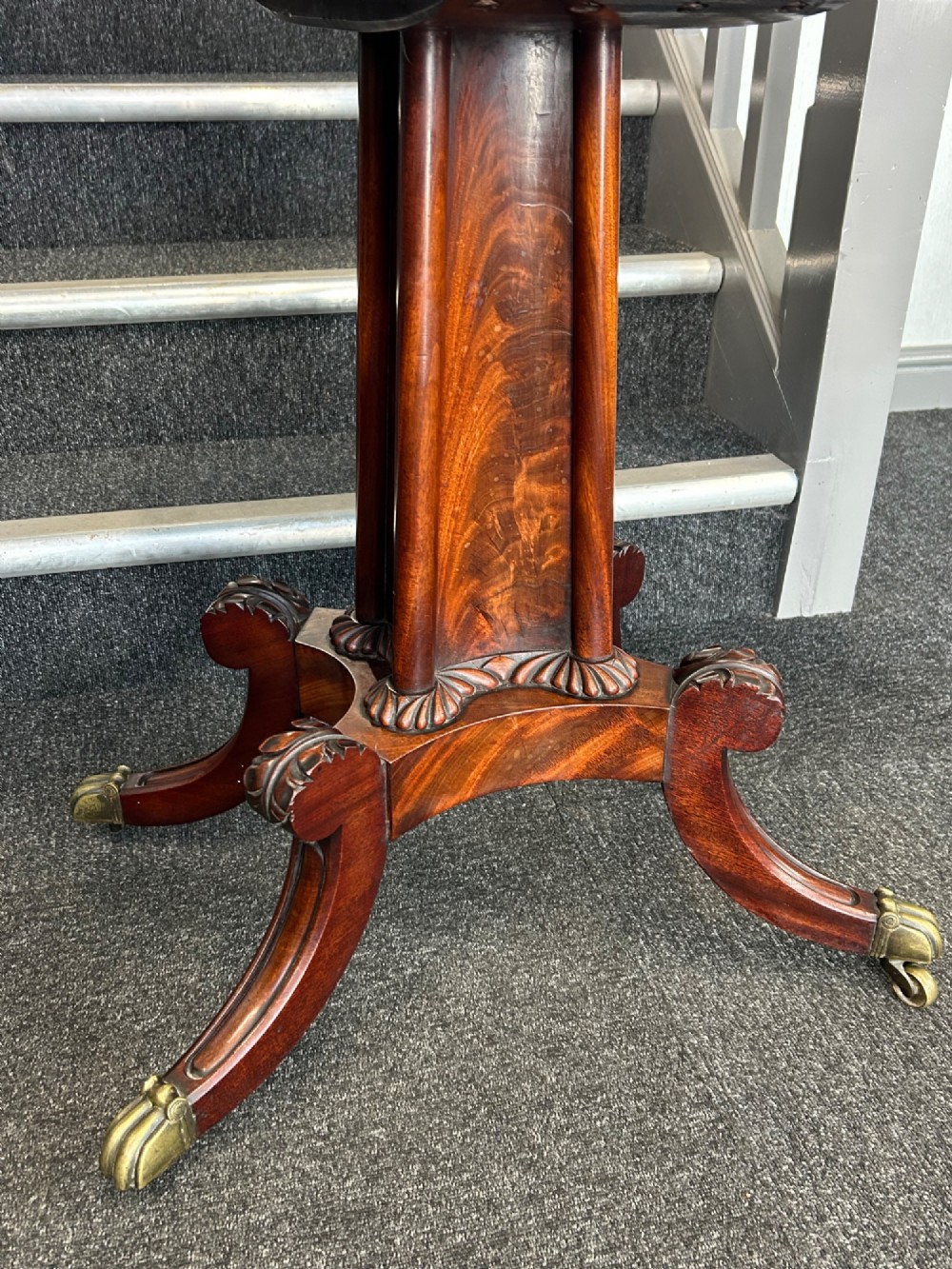 A William IV mahogany fold-over tea table with a flame mahogany top, central carved pedestal, and four splayed legs with brass paw castors. View II