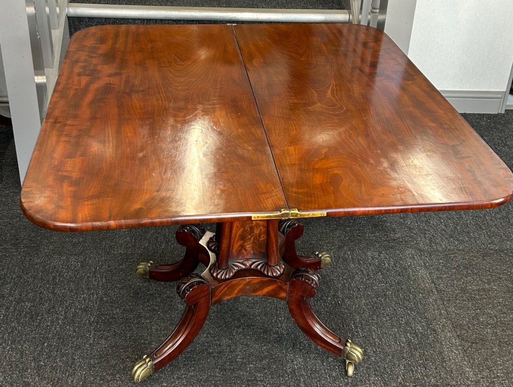 A William IV mahogany fold-over tea table with a flame mahogany top, central carved pedestal, and four splayed legs with brass paw castors. View III