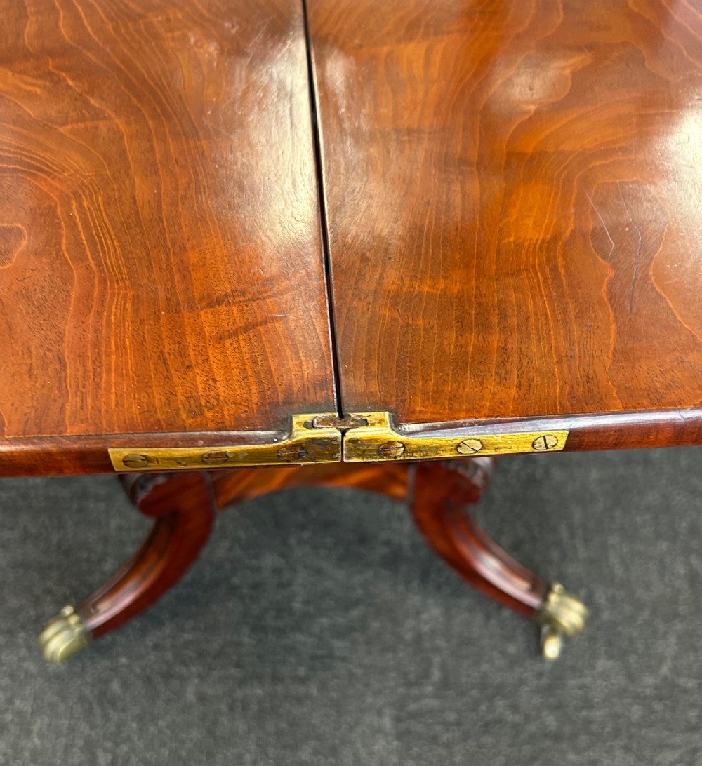 A William IV mahogany fold-over tea table with a flame mahogany top, central carved pedestal, and four splayed legs with brass paw castors. View IV