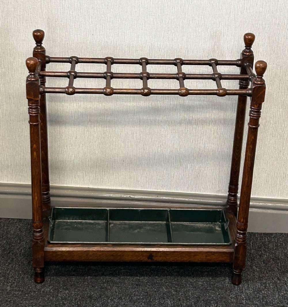 A dark oak stick stand featuring a multi-section top grid, four turned corner posts with finials, and a removable dark metal drip tray.