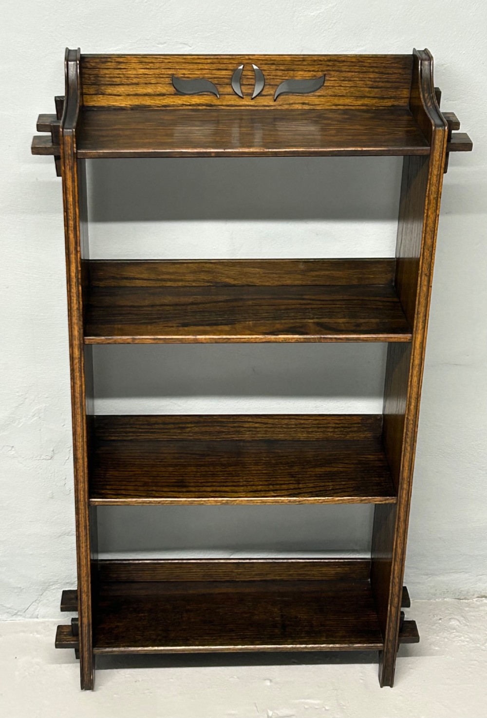A small Arts and Crafts dark solid oak open bookcase with four shelves, featuring extended side tenons and a stylised pierced cutout motif on the top gallery.
