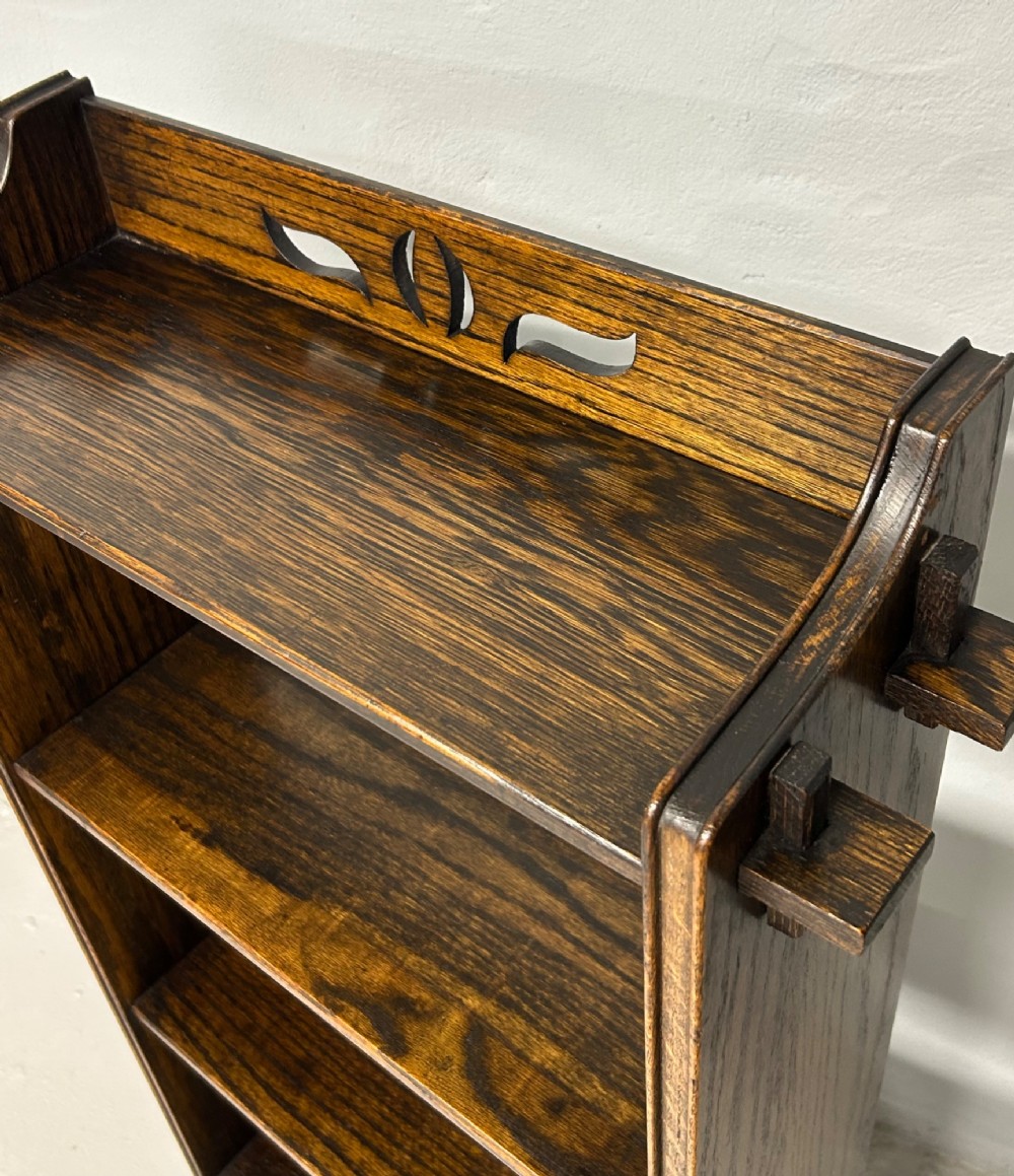 A small Arts and Crafts dark solid oak open bookcase with four shelves, featuring extended side tenons and a stylised pierced cutout motif on the top gallery. View III