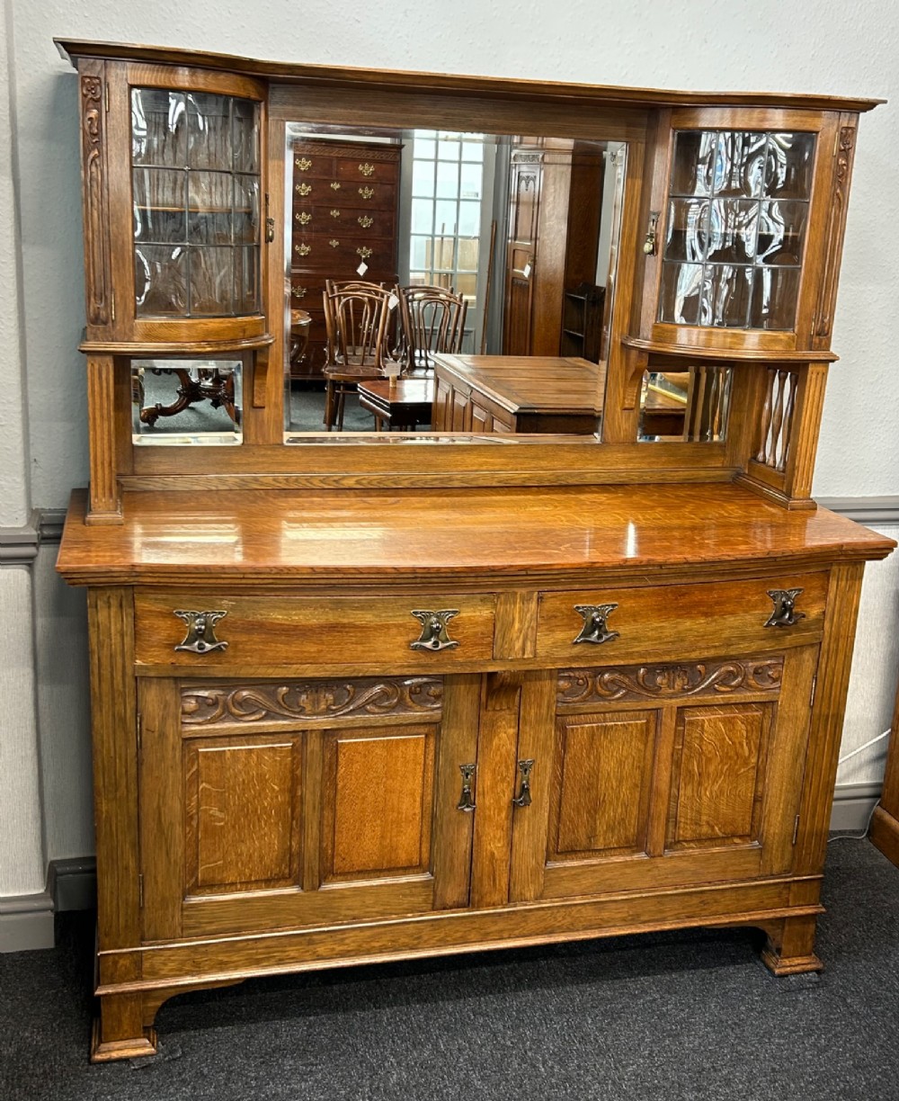 An Edwardian Arts and Crafts oak sideboard featuring a central mirrored superstructure flanked by bow-fronted leaded glass cabinets above drawers and carved panel doors.