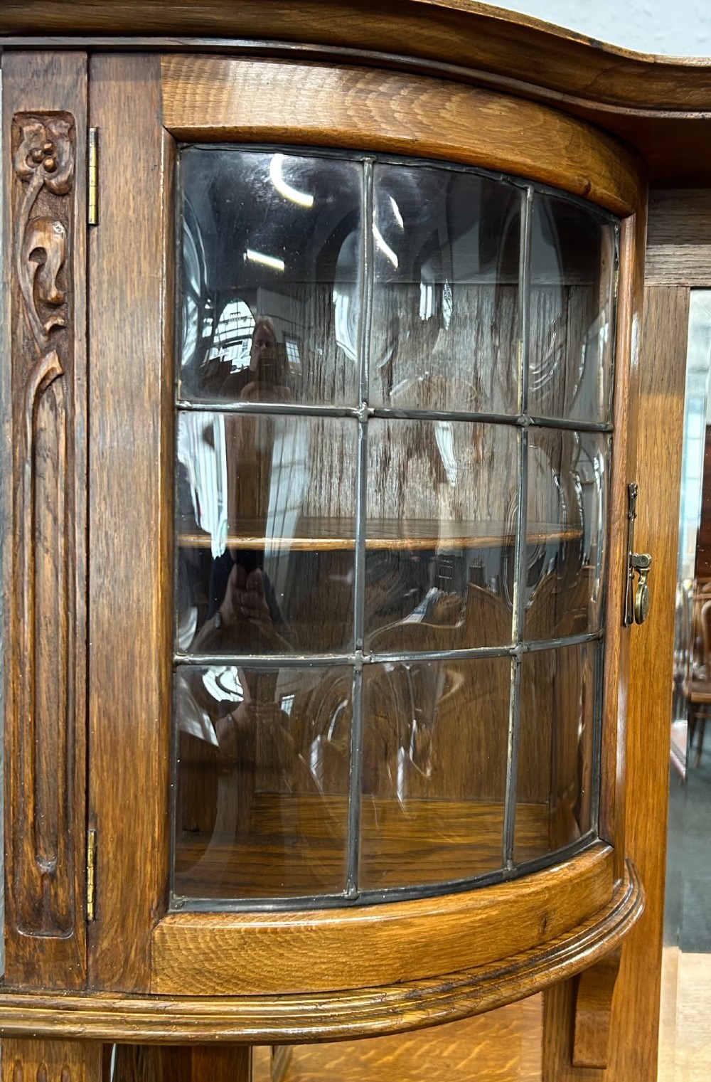 An Edwardian Arts and Crafts oak sideboard featuring a central mirrored superstructure flanked by bow-fronted leaded glass cabinets above drawers and carved panel doors. View II