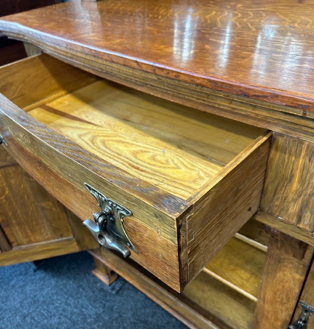 An Edwardian Arts and Crafts oak sideboard featuring a central mirrored superstructure flanked by bow-fronted leaded glass cabinets above drawers and carved panel doors. View X