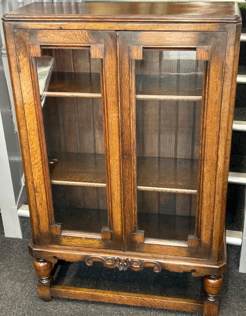 A medium brown oak cabinet featuring two glass-panelled doors, internal wooden shelves, a carved decorative apron, and turned legs with connecting stretchers.