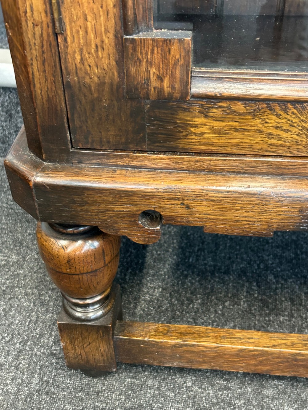 A medium brown oak cabinet featuring two glass-panelled doors, internal wooden shelves, a carved decorative apron, and turned legs with connecting stretchers. View III