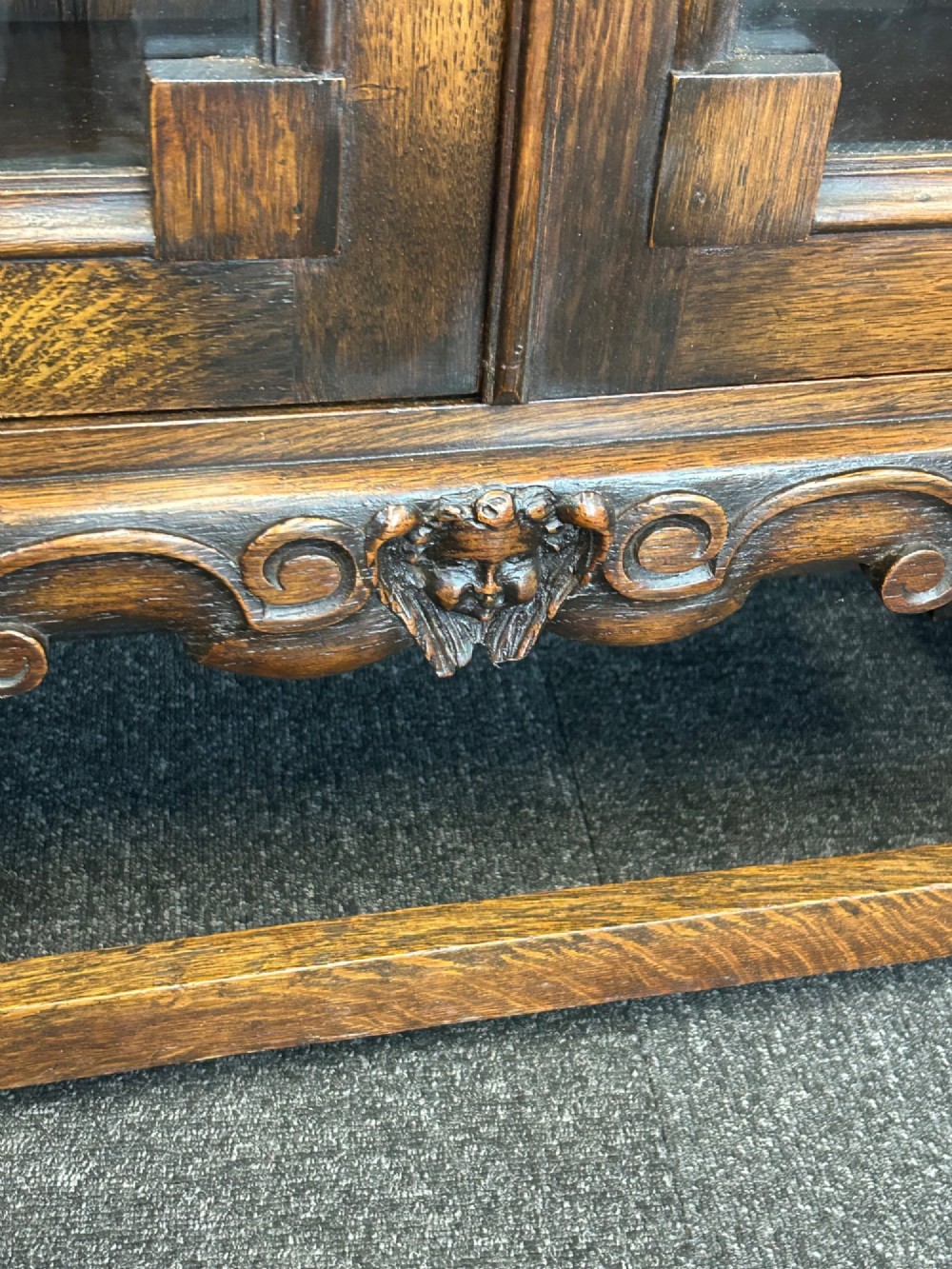 A medium brown oak cabinet featuring two glass-panelled doors, internal wooden shelves, a carved decorative apron, and turned legs with connecting stretchers. View IV