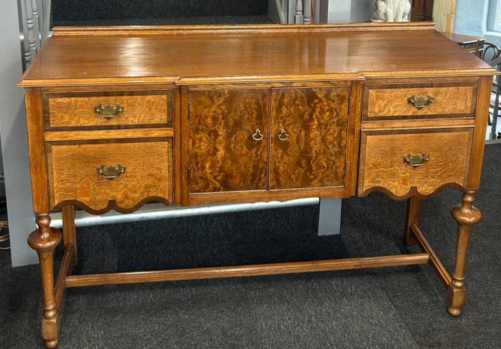 An antique oak sideboard featuring central burr walnut cupboard doors, four side drawers, brass drop handles, shaped aprons, and turned legs with a stretcher base.