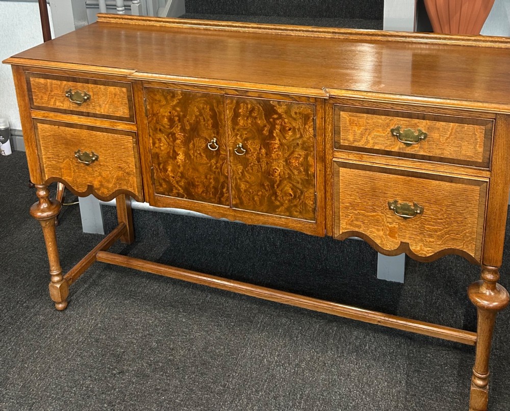 An antique oak sideboard featuring central burr walnut cupboard doors, four side drawers, brass drop handles, shaped aprons, and turned legs with a stretcher base. View II