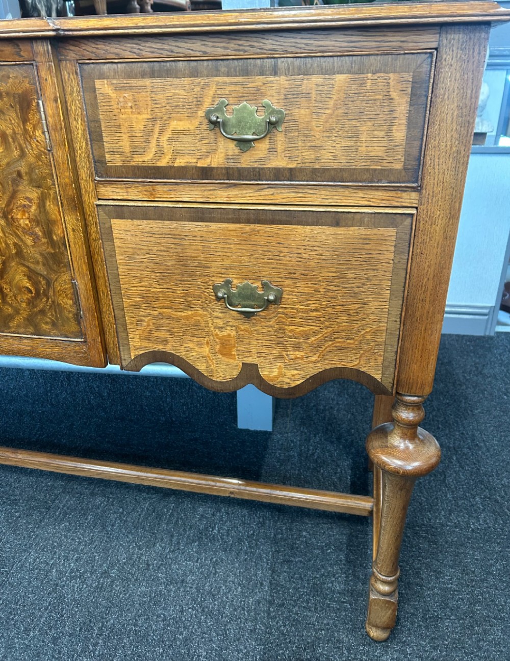 An antique oak sideboard featuring central burr walnut cupboard doors, four side drawers, brass drop handles, shaped aprons, and turned legs with a stretcher base. View IV