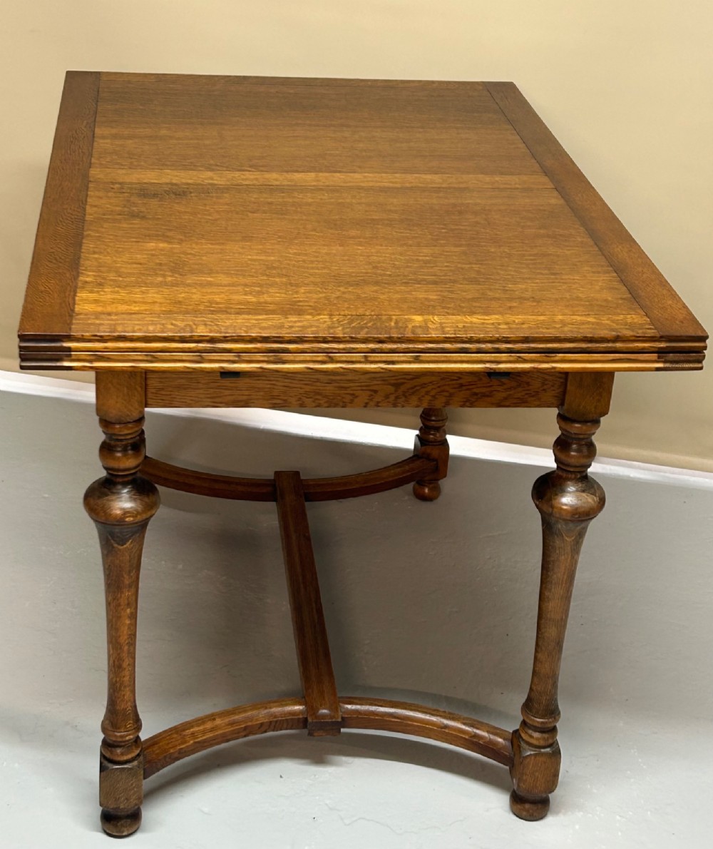 A medium brown oak draw-leaf extending dining table featuring turned baluster legs joined by a curved stretcher base.