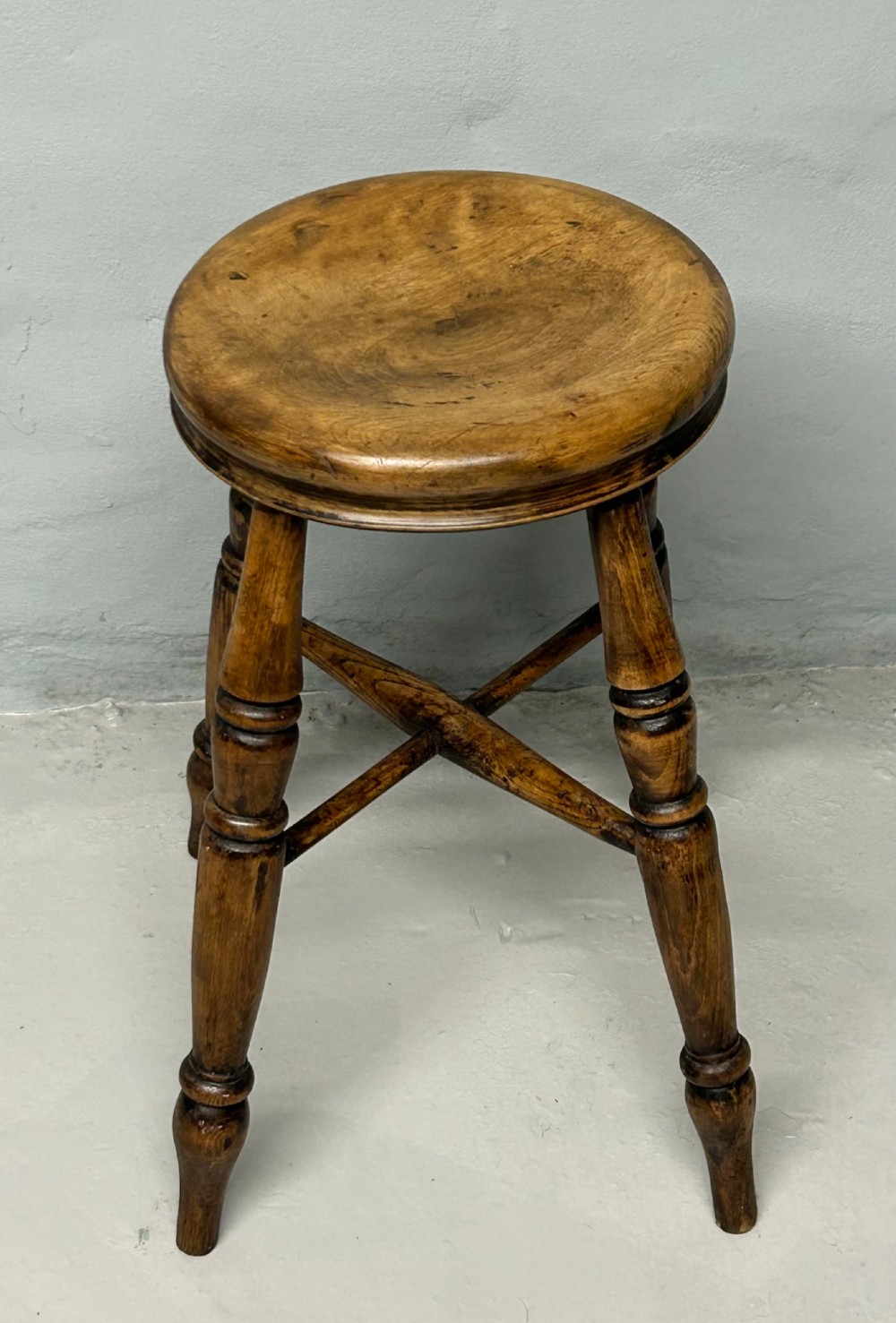 An antique wooden tavern stool with a circular dished elm seat, four turned beech legs, and cross stretchers in a warm brown timber finish.