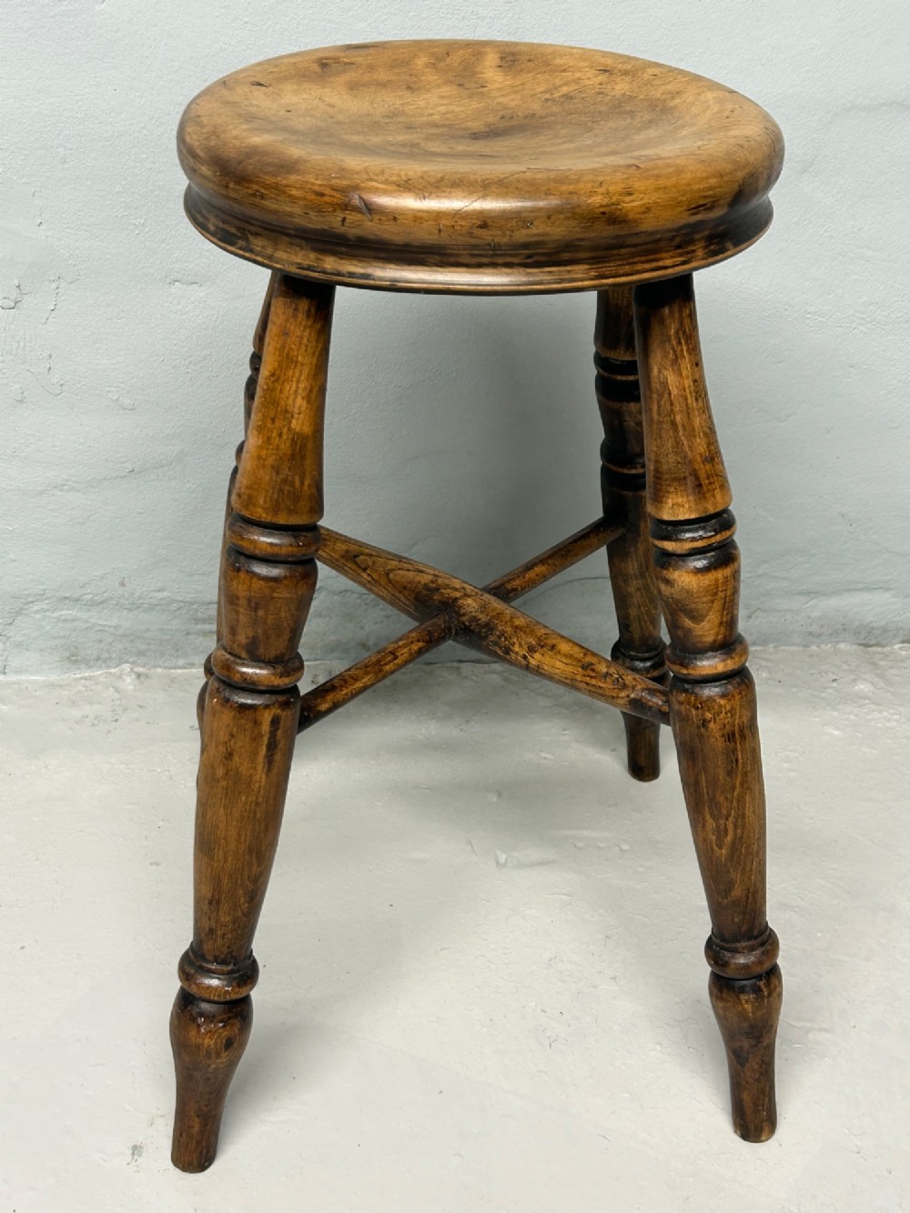 An antique wooden tavern stool with a circular dished elm seat, four turned beech legs, and cross stretchers in a warm brown timber finish. View II