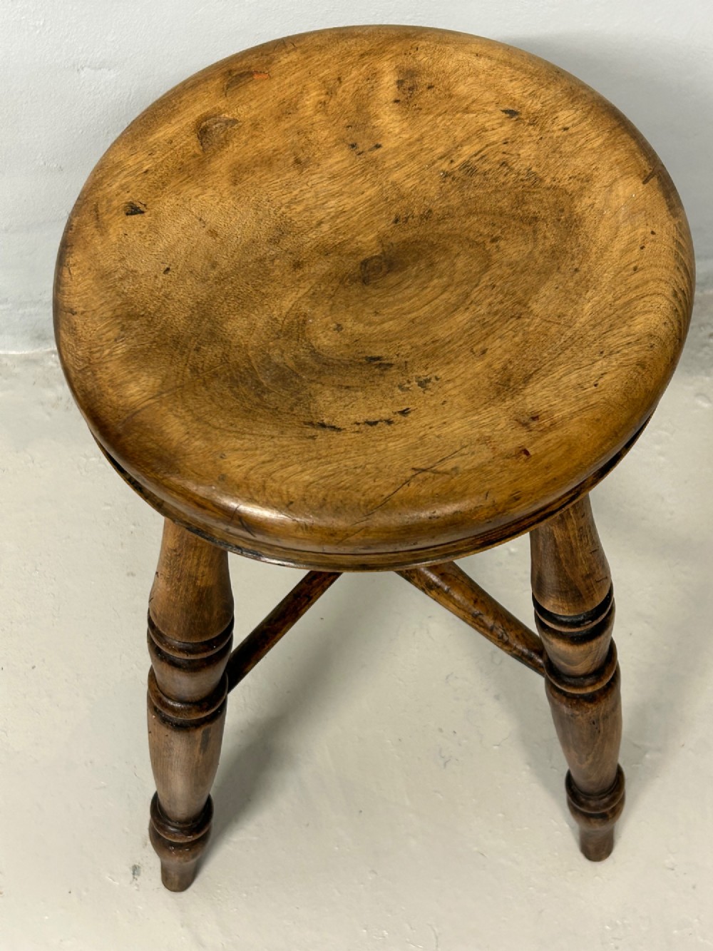 An antique wooden tavern stool with a circular dished elm seat, four turned beech legs, and cross stretchers in a warm brown timber finish. View III