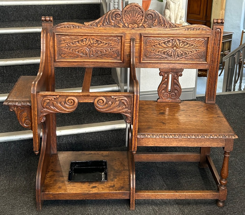 An antique carved oak hall seat featuring an integrated umbrella stand with drip tray, side shelf, and carved foliate back panels topped with a shell pediment.