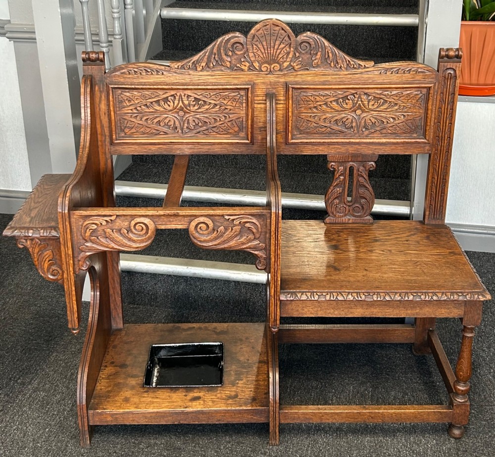 An antique carved oak hall seat featuring an integrated umbrella stand with drip tray, side shelf, and carved foliate back panels topped with a shell pediment. View II