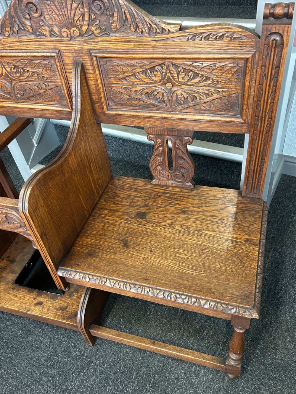 An antique carved oak hall seat featuring an integrated umbrella stand with drip tray, side shelf, and carved foliate back panels topped with a shell pediment. View V