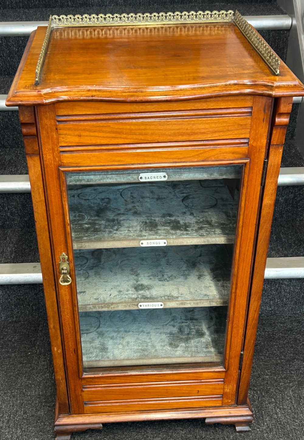 An Edwardian walnut music cabinet featuring a brass gallery top rail, a single glazed door, and lined interior shelves with small brass labels. View III