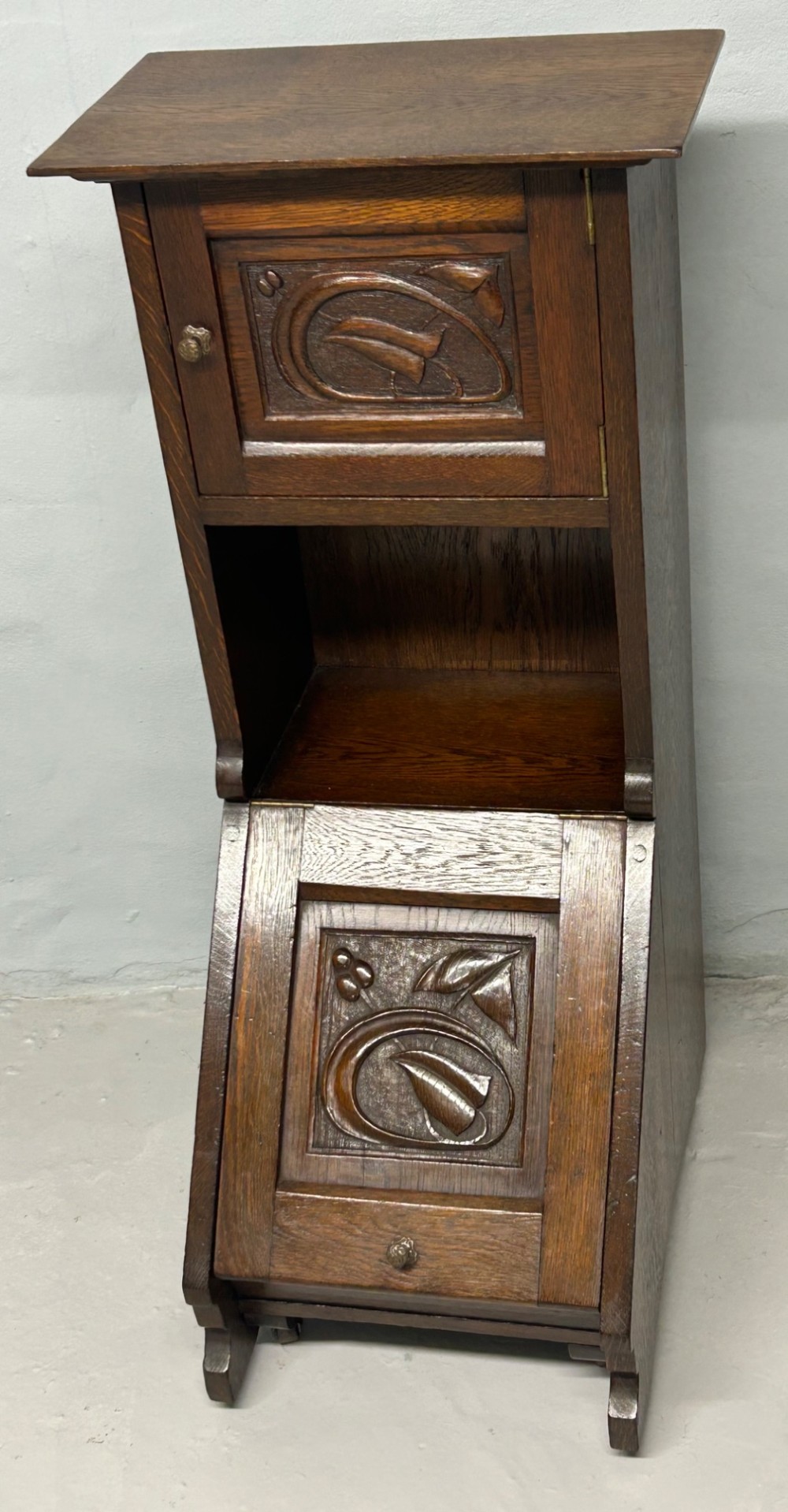 An Arts and Crafts dark oak purdonium featuring stylized carved leaf panels, an upper cupboard, an open shelf, and a slanted fall-front compartment. View II