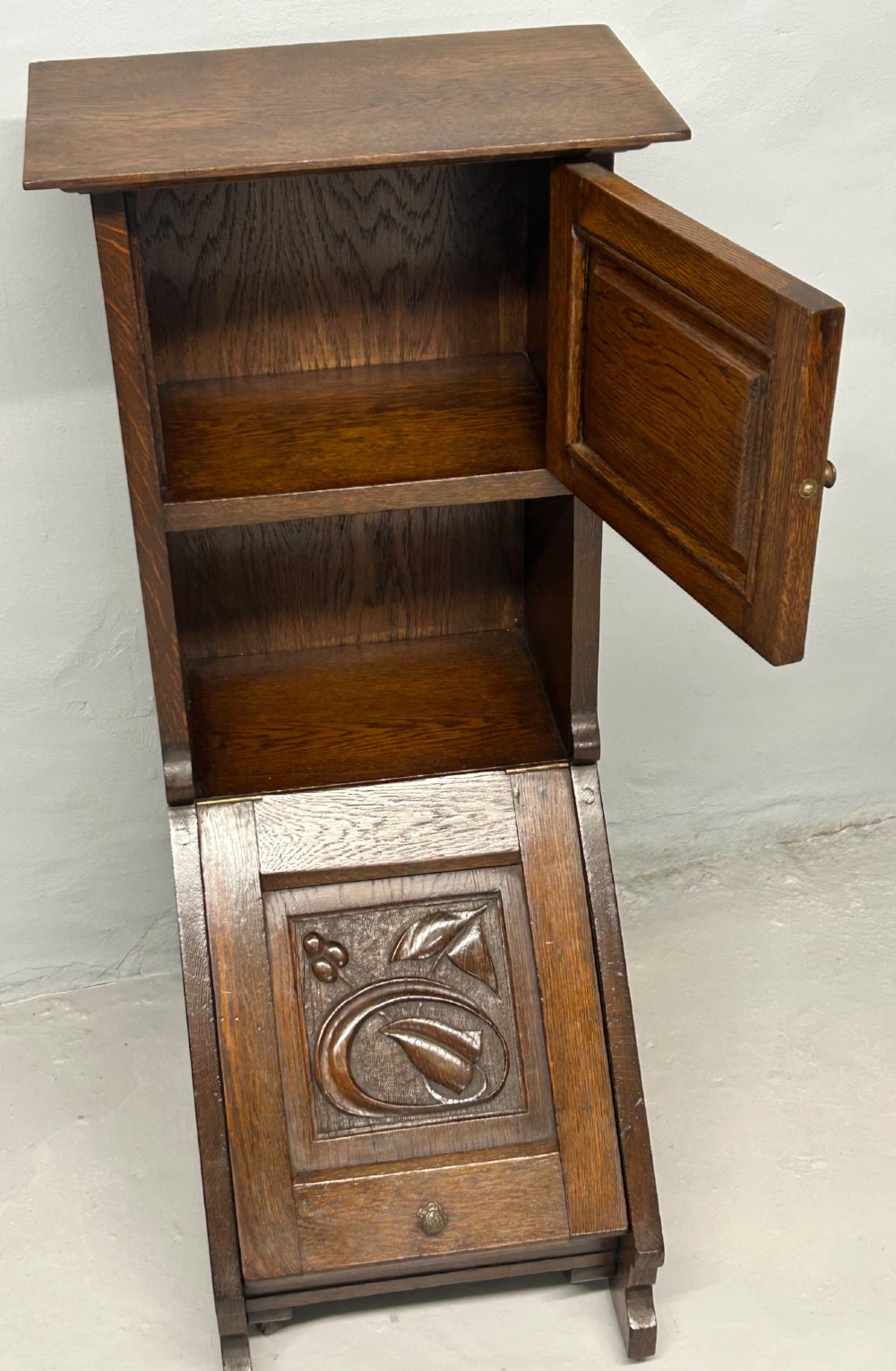 An Arts and Crafts dark oak purdonium featuring stylized carved leaf panels, an upper cupboard, an open shelf, and a slanted fall-front compartment. View III