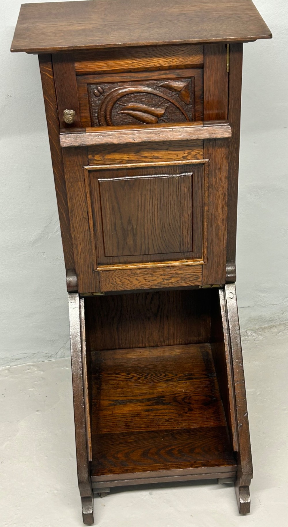 An Arts and Crafts dark oak purdonium featuring stylized carved leaf panels, an upper cupboard, an open shelf, and a slanted fall-front compartment. View IV