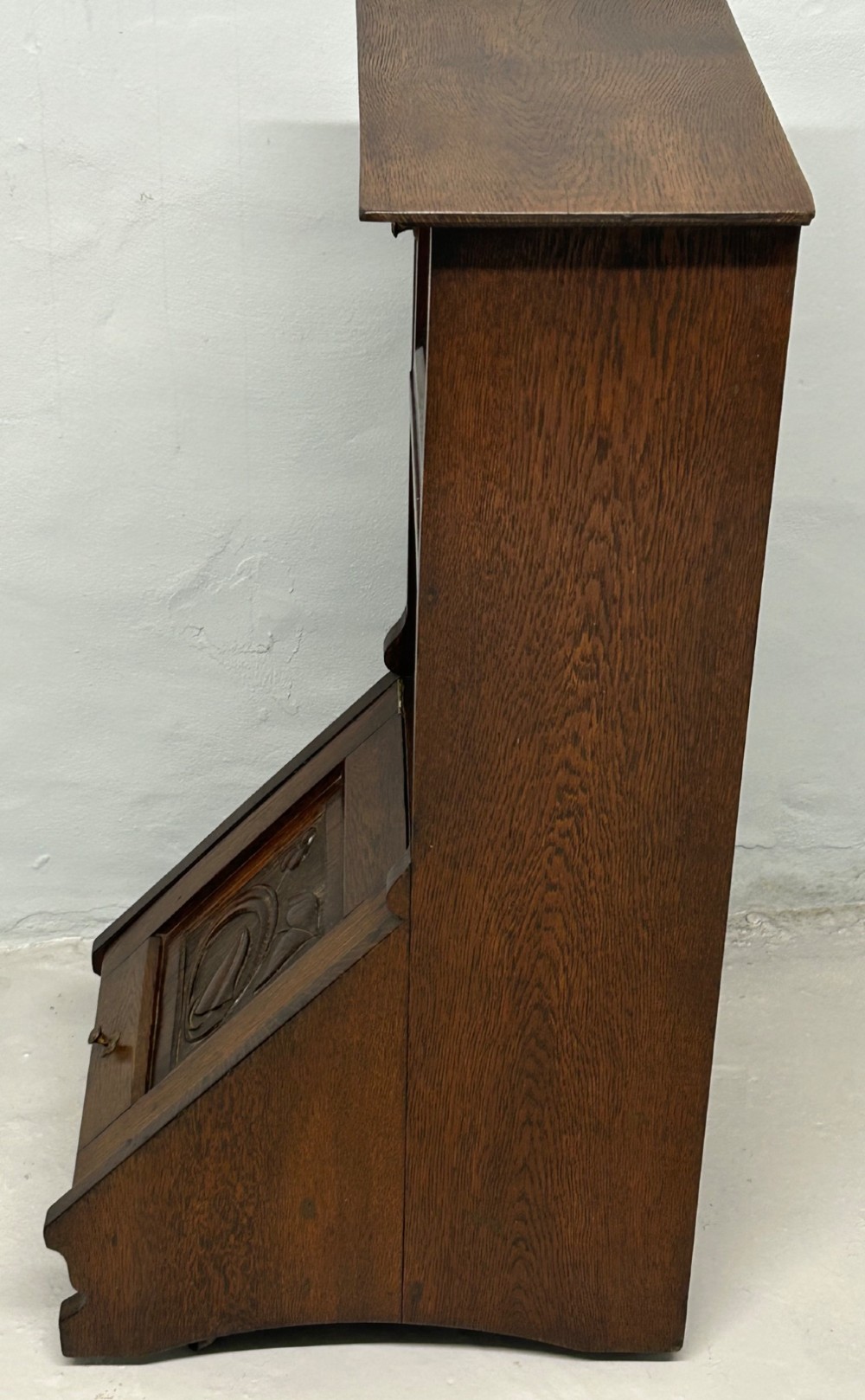 An Arts and Crafts dark oak purdonium featuring stylized carved leaf panels, an upper cupboard, an open shelf, and a slanted fall-front compartment. View V