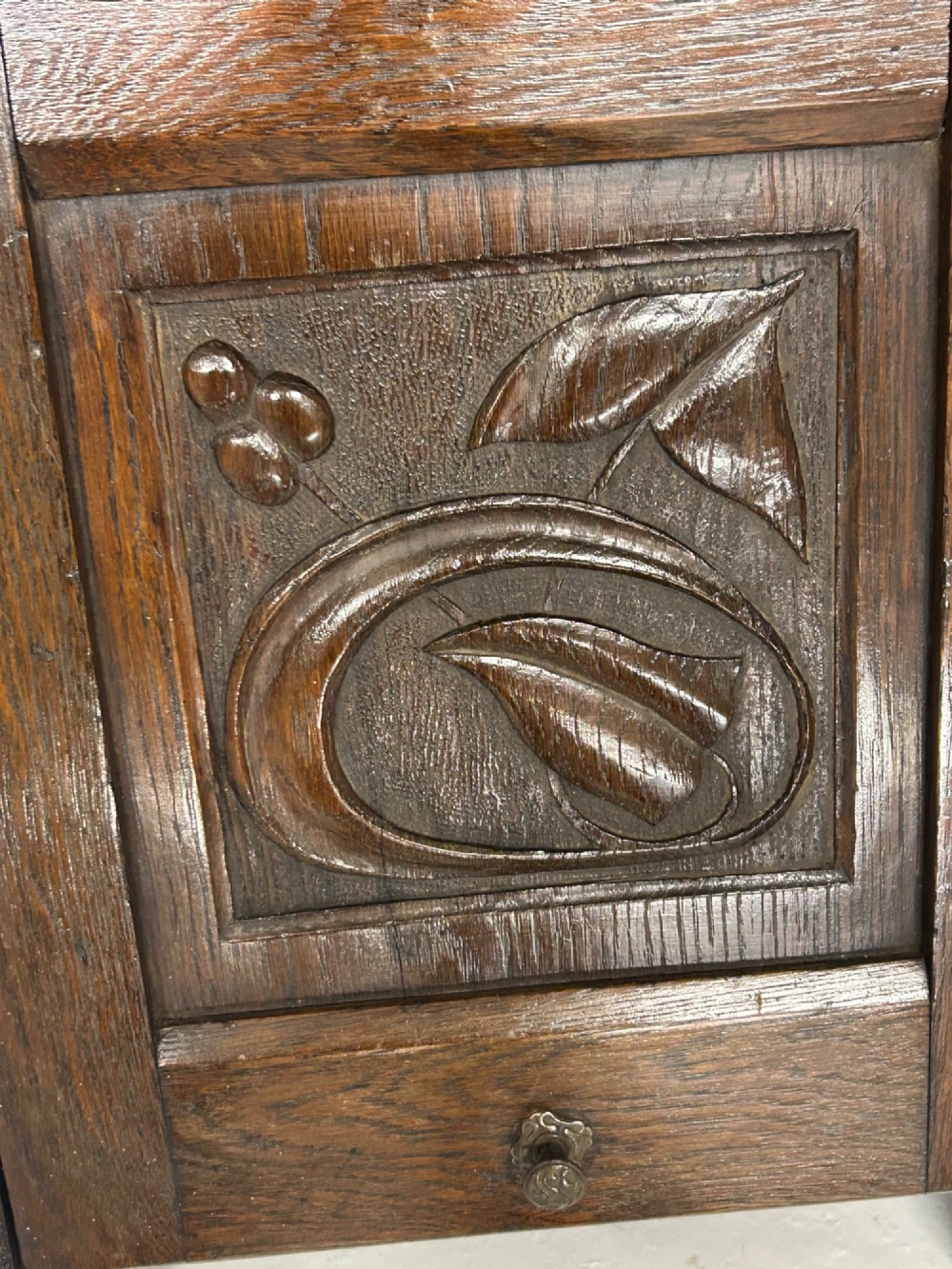 An Arts and Crafts dark oak purdonium featuring stylized carved leaf panels, an upper cupboard, an open shelf, and a slanted fall-front compartment. View VII
